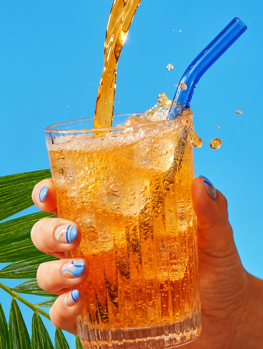 La bebida servíndose en un vaso con hielo sobre fondo azul, junto a una hoja de palma